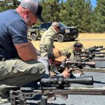 fundamentalsLevel2SomeFreeWisdomGallery6 Four men inspect rifles arranged in a row on a shooting platform.